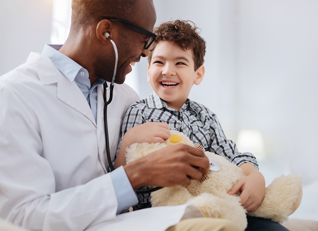 Insurance by Industry - Portrait of a Cheerful Young Male Doctor with a Young Boy Giving a Teddy Bear a Medical Exam in an Office